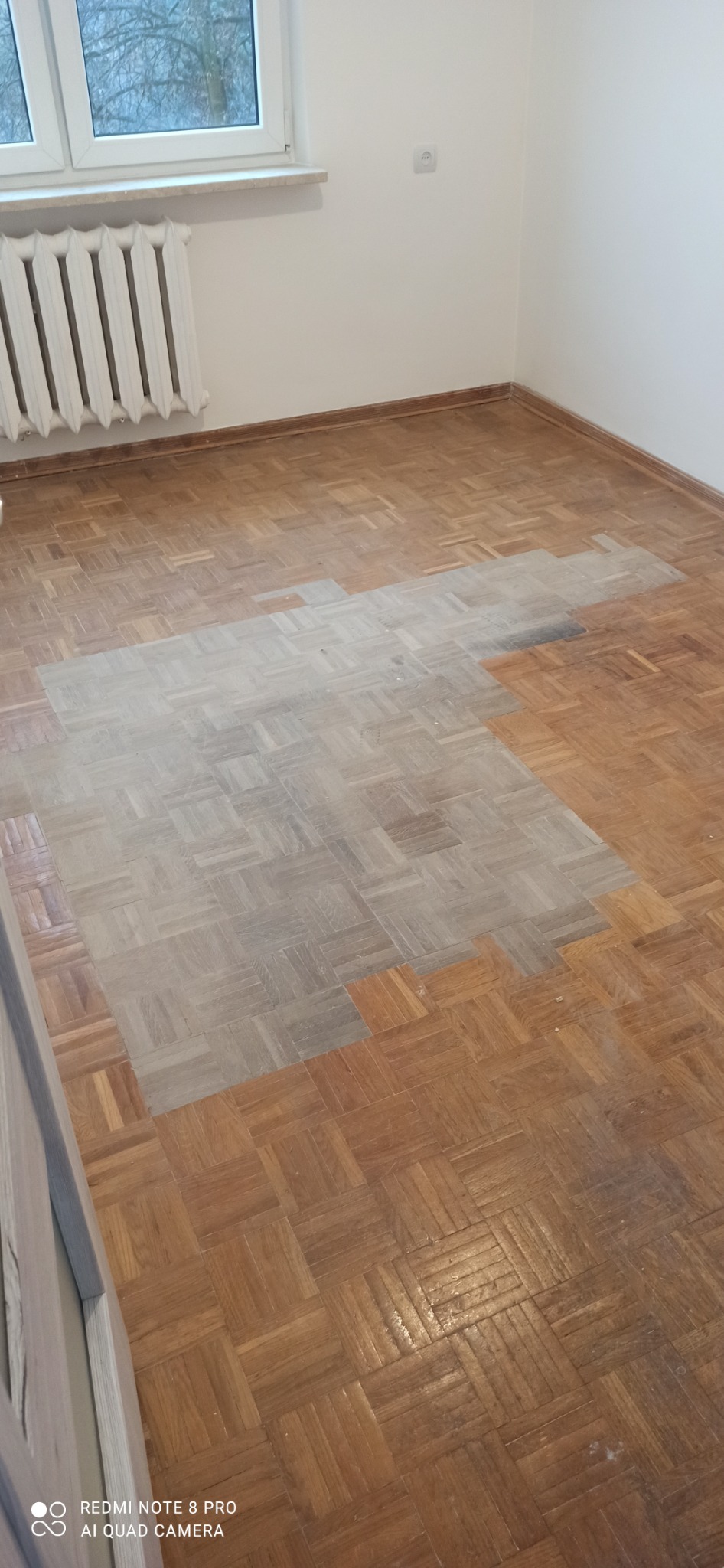 Częściowo wycyklinowany parkiet w jodełkę, widoczna różnica w kolorze i fakturze między obszarem po obróbce a nieobrobionym, w tle biały grzejnik i okno.