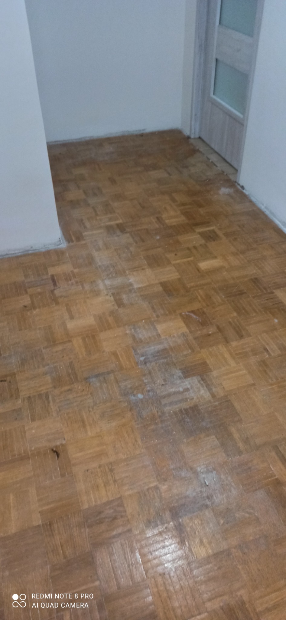 Parkiet ułożony w kwadraty, częściowo pokryty białym pyłem, widoczne ślady po szpachlowaniu lub cyklinowaniu, ściany białe, w tle drzwi.