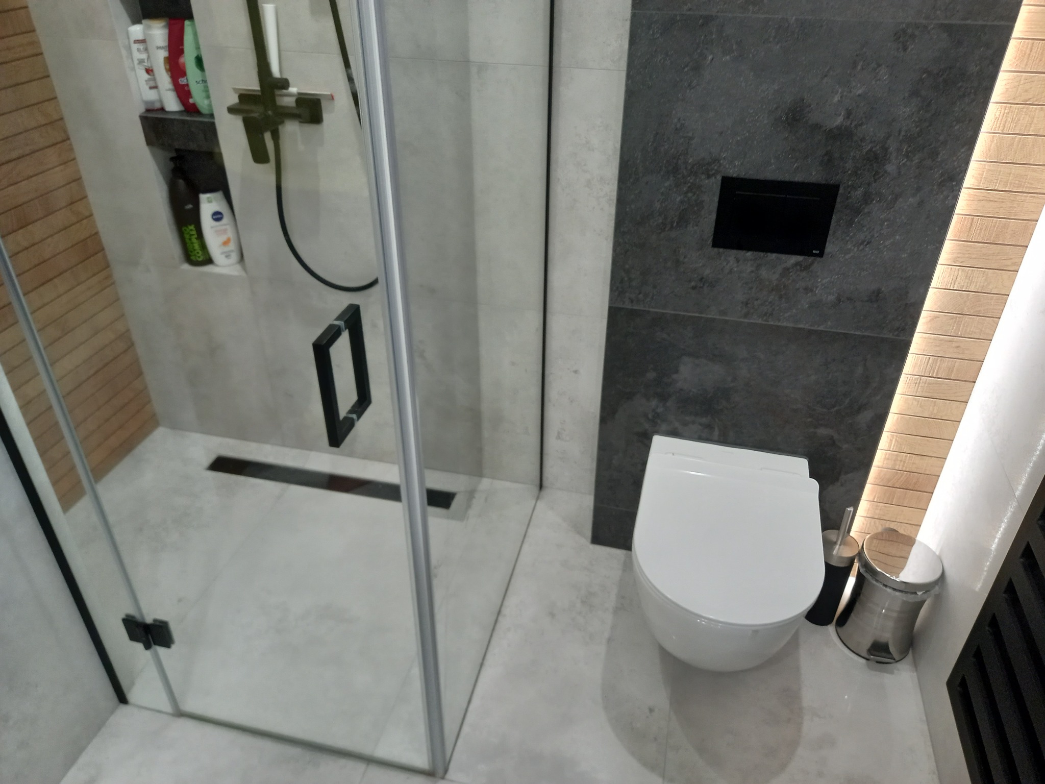 Nowoczesna łazienka z kabiną prysznicową ze szklanymi drzwiami, odpływem liniowym i wiszącą toaletą z szarą płytką imitującą kamień oraz drewnianymi panelami na ścianie.