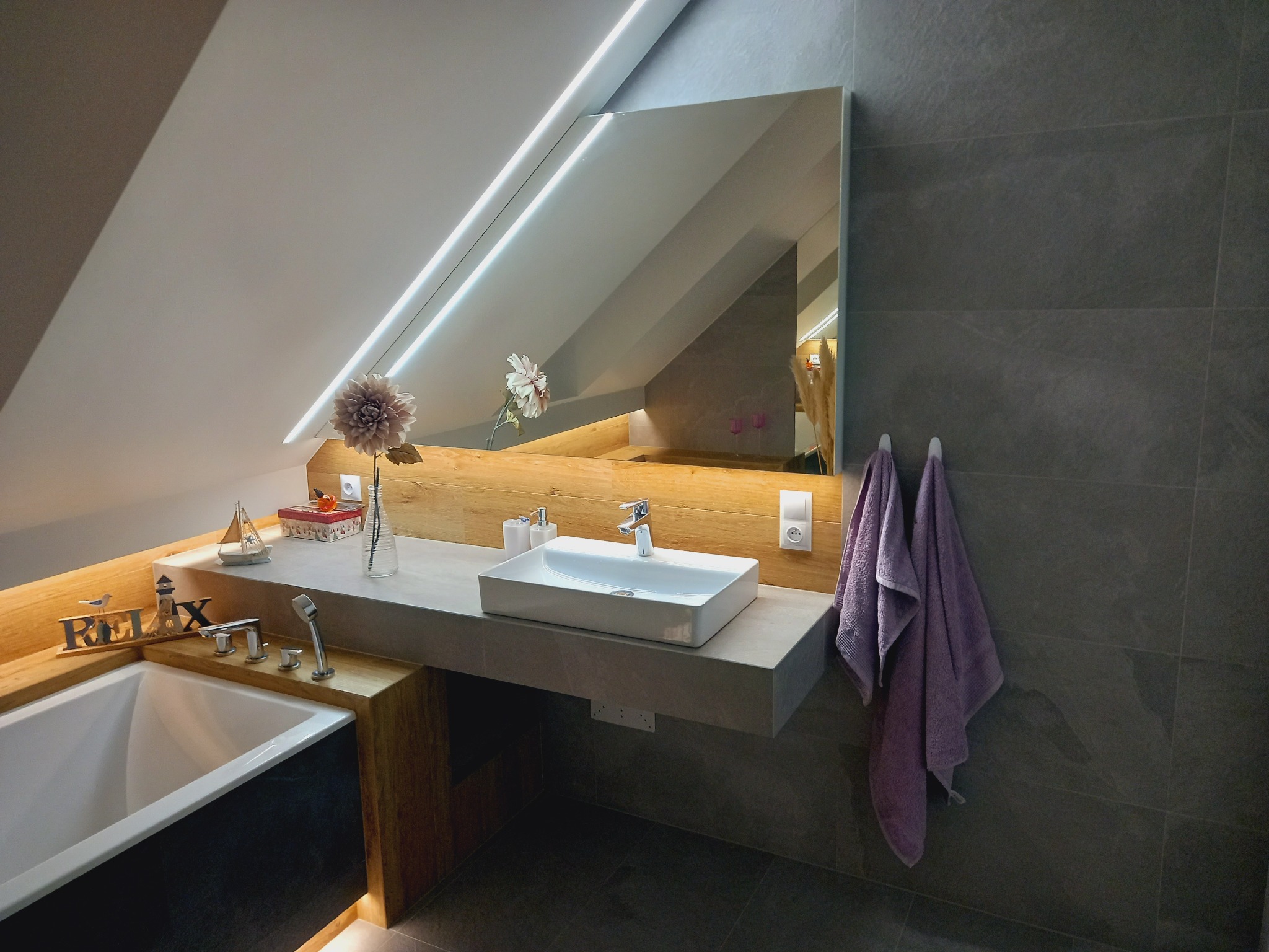 Nowoczesna łazienka w Osobnicy z szarą glazurą, podwieszaną umywalką, lustrem z oświetleniem LED i dekoracyjnymi elementami, takimi jak kwiaty i figurki.