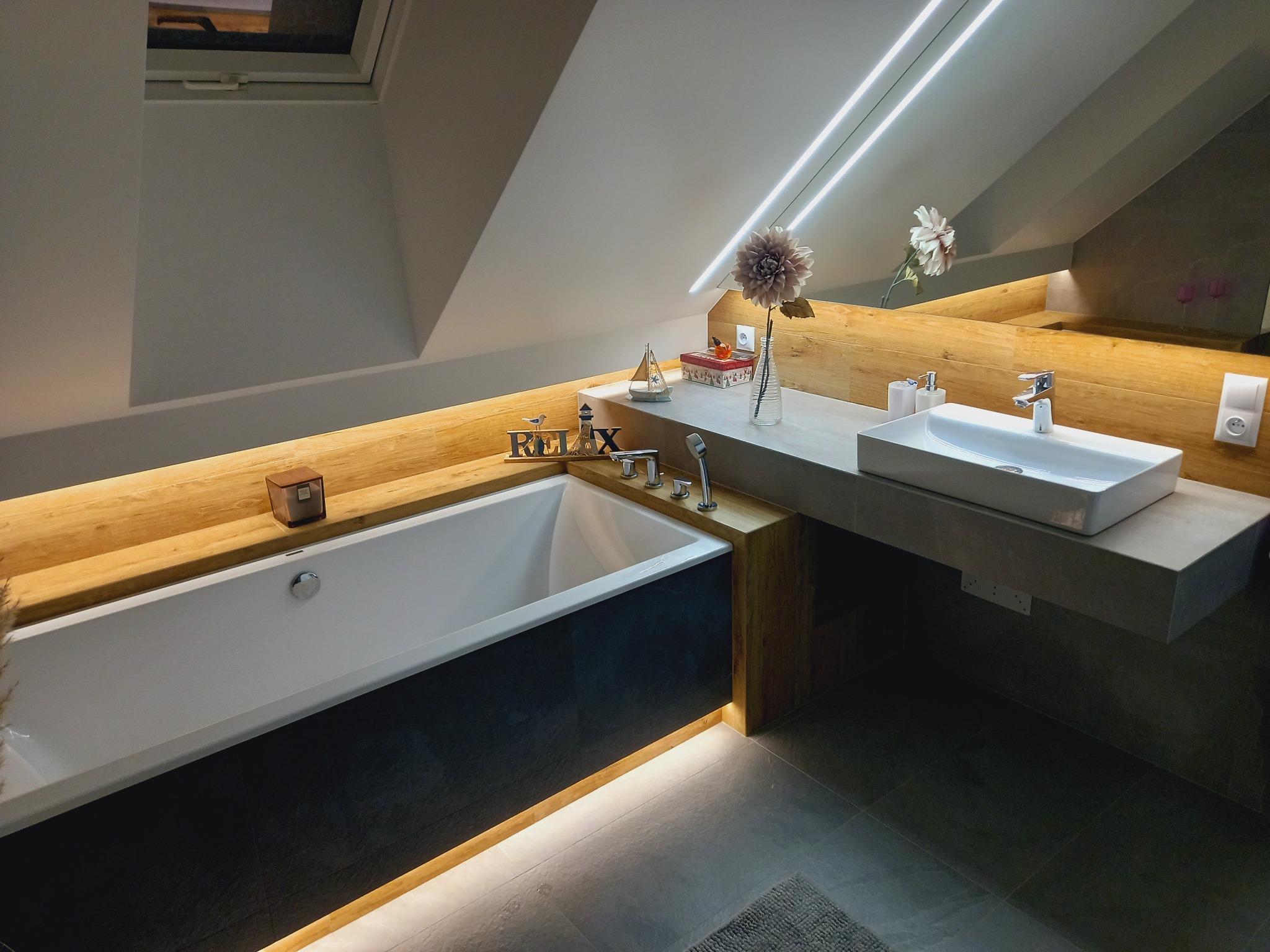 Nowoczesna łazienka na poddaszu z wanną obudowaną ciemną płytą, drewnianym blatem, umywalką nablatową i dekoracyjnym oświetleniem LED.