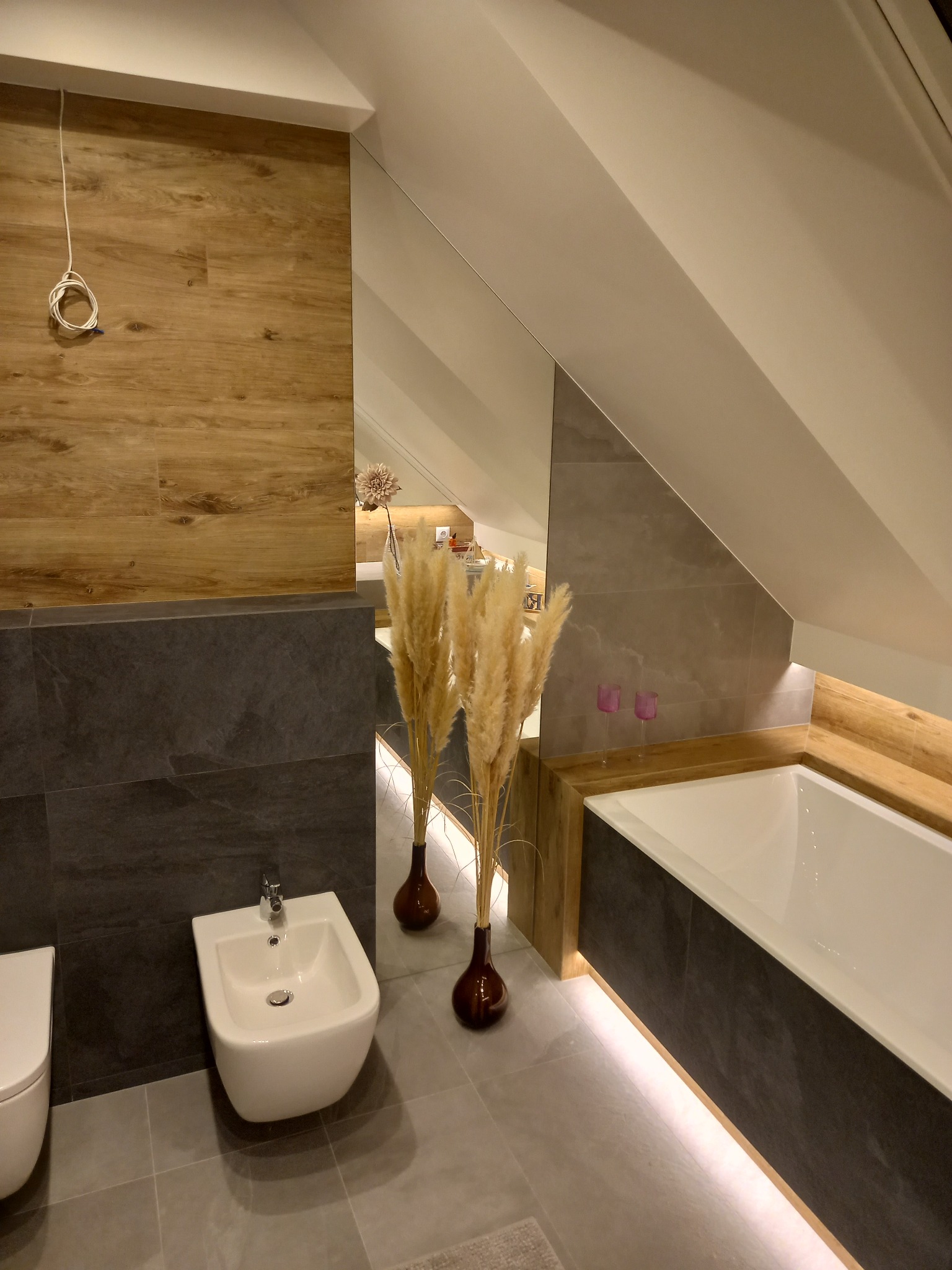 Nowoczesna łazienka na poddaszu z drewnopodobnymi i grafitowymi płytkami, białą armaturą, wanną z podświetleniem LED oraz dekoracyjnymi trawami pampasowymi w wazonach.