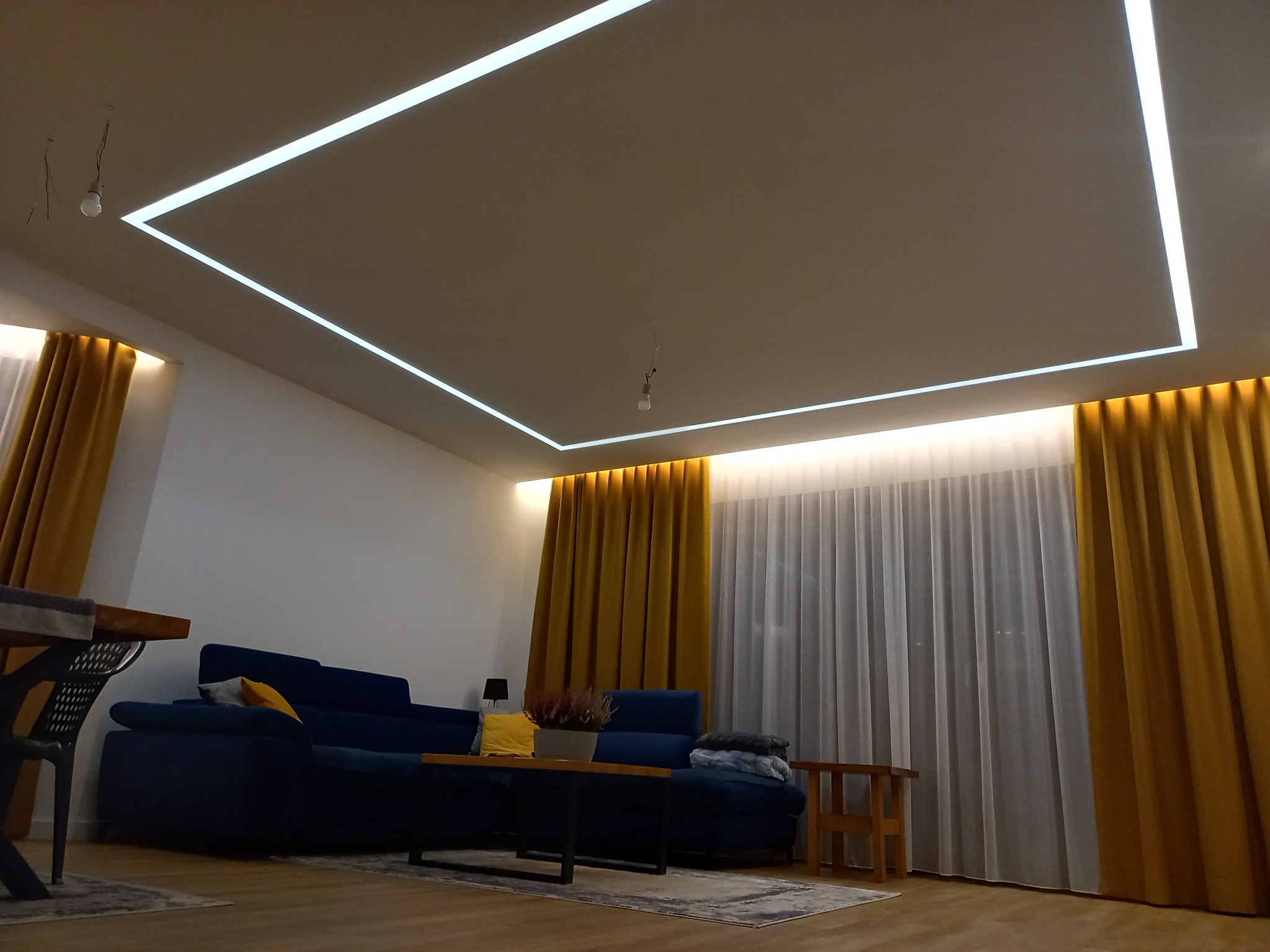 Nowoczesny salon z podwieszonym sufitem z oświetleniem LED w kształcie prostokąta, niebieska sofa z żółtymi poduszkami, drewniany stół i złote zasłony w tle.