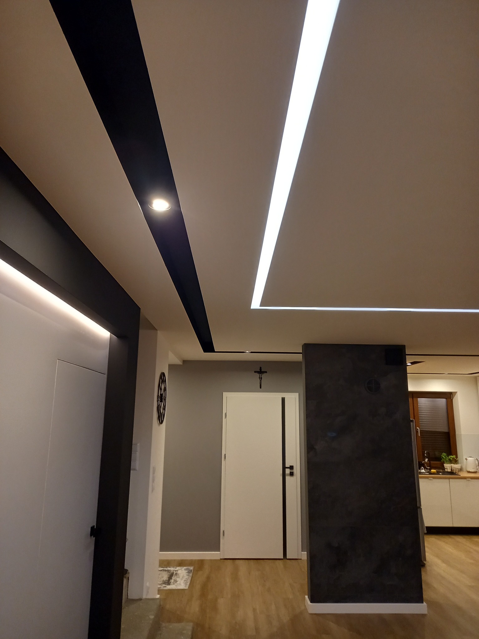 Nowoczesne wnętrze mieszkania z podwieszanym sufitem z oświetleniem LED i czarnymi akcentami, widok na korytarz i częściowo na kuchnię.