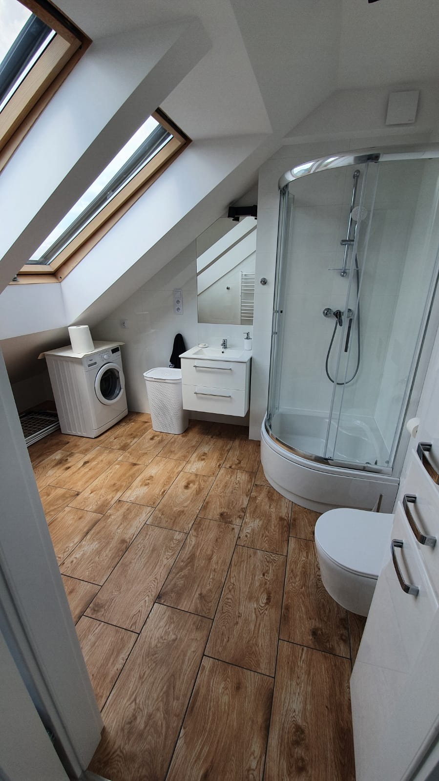 Jasna łazienka na poddaszu z drewnopodobną podłogą, kabiną prysznicową, umywalką z szafką, toaletą i pralką pod oknem dachowym.