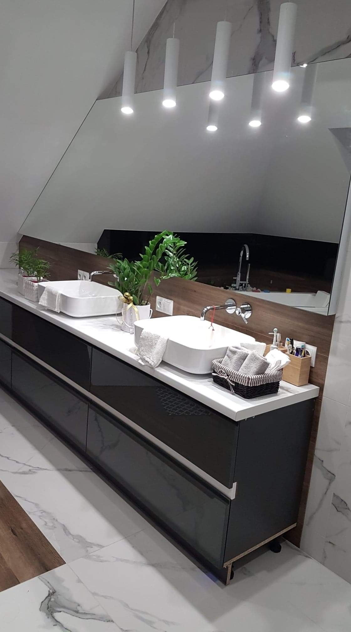 Nowoczesna łazienka z dwoma białymi umywalkami nablatowymi, ciemnymi szafkami z połyskiem, dużym lustrem, roślinami doniczkowymi i marmurową podłogą.