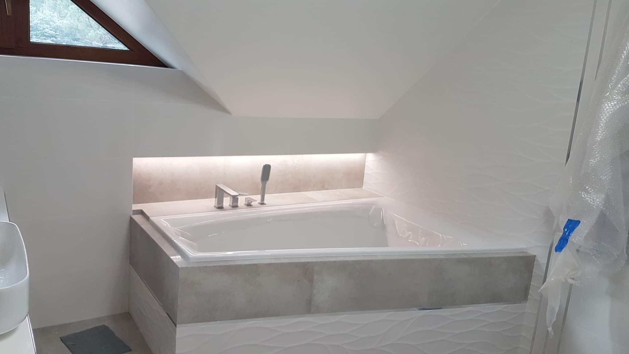 Nowoczesna łazienka na poddaszu: wanna obudowana szarymi płytkami, białe ściany z delikatnym wzorem, oświetlenie LED nad wanną, bateria wannowa w minimalistycznym stylu, okno dachowe.