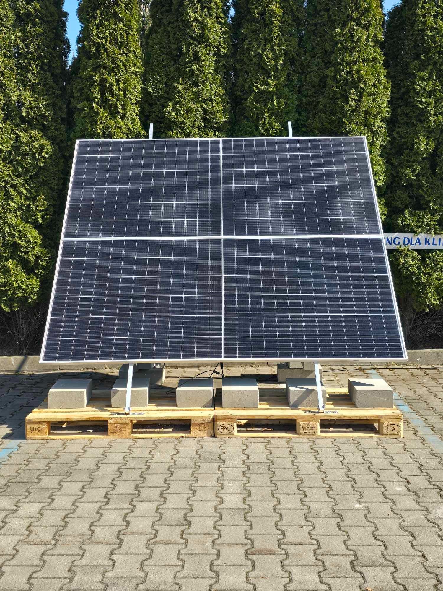 Cztery panele fotowoltaiczne ustawione na betonowych blokach i paletach euro, z zielonymi tujami w tle i kostką brukową na pierwszym planie.