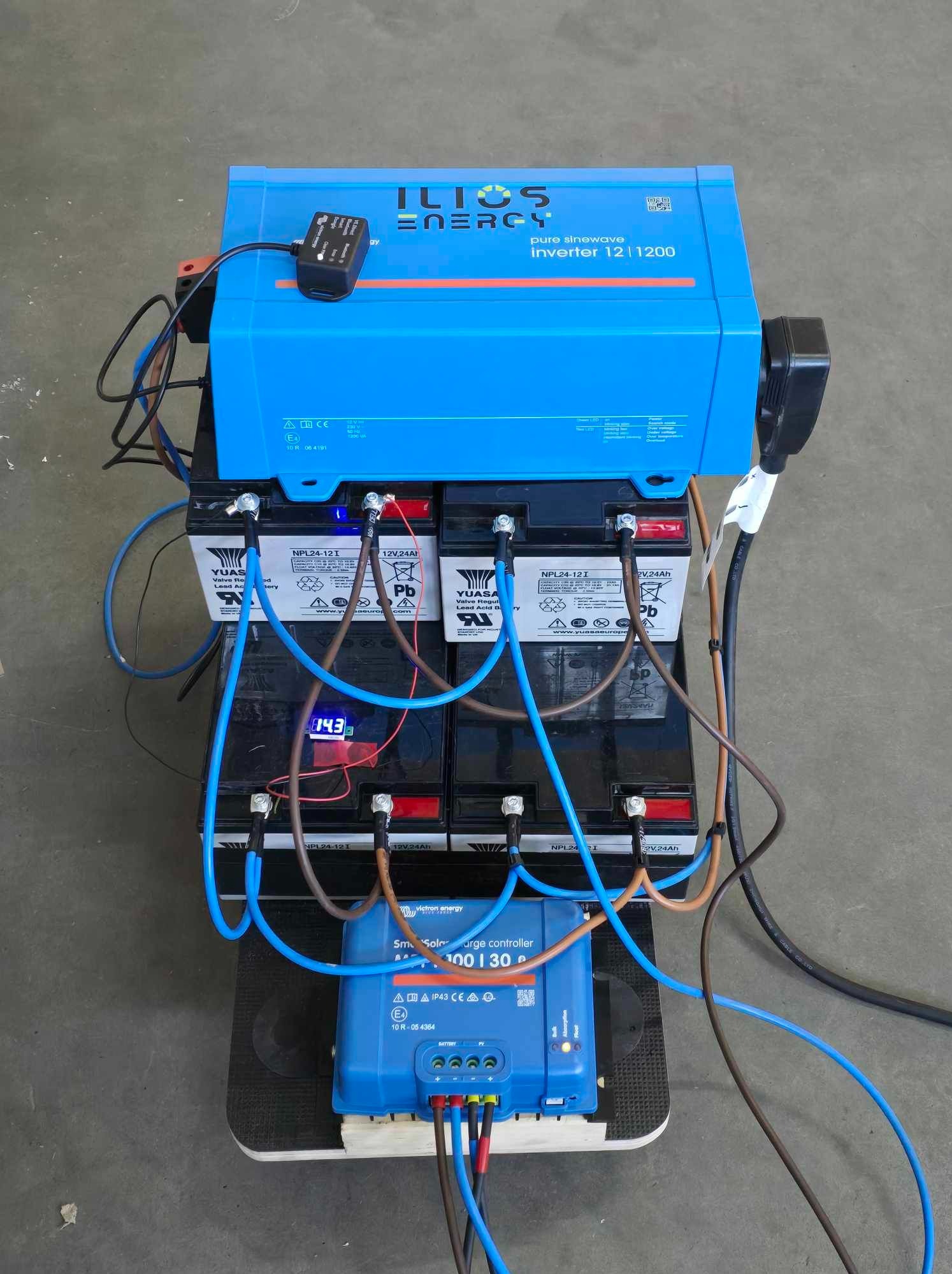 Zestaw akumulatorów 12V, inwerter ILIOS Energy 12/1200 i regulator ładowania Victron Energy SmartSolar MPPT 100/30, połączone przewodami na szarej podłodze.