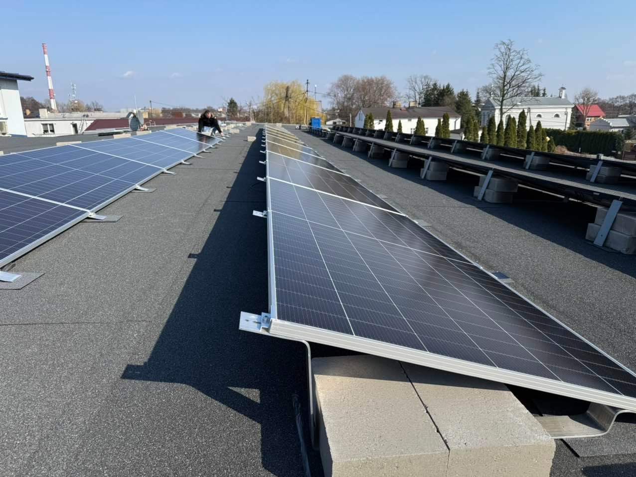 Instalacja paneli słonecznych na płaskim dachu z widocznymi betonowymi podstawami, w tle budynki mieszkalne i komin