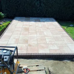 Świeżo ułożony plac z beżowej kostki brukowej otoczony ciemniejszą obwódką, z ubijarką wibracyjną i narzędziami na palecie w pierwszym planie, a w tle gęsty żywopłot.