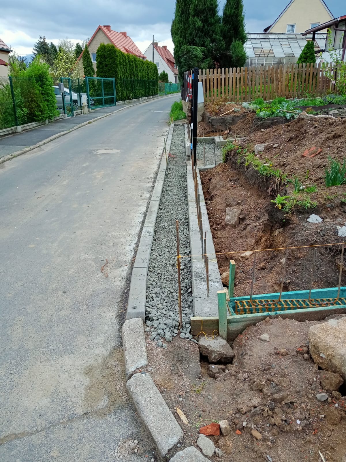 Przygotowanie podłoża pod obrzeże chodnika z wysypanym grysem, widoczne szalunki i pręty zbrojeniowe, w tle ulica i domy.