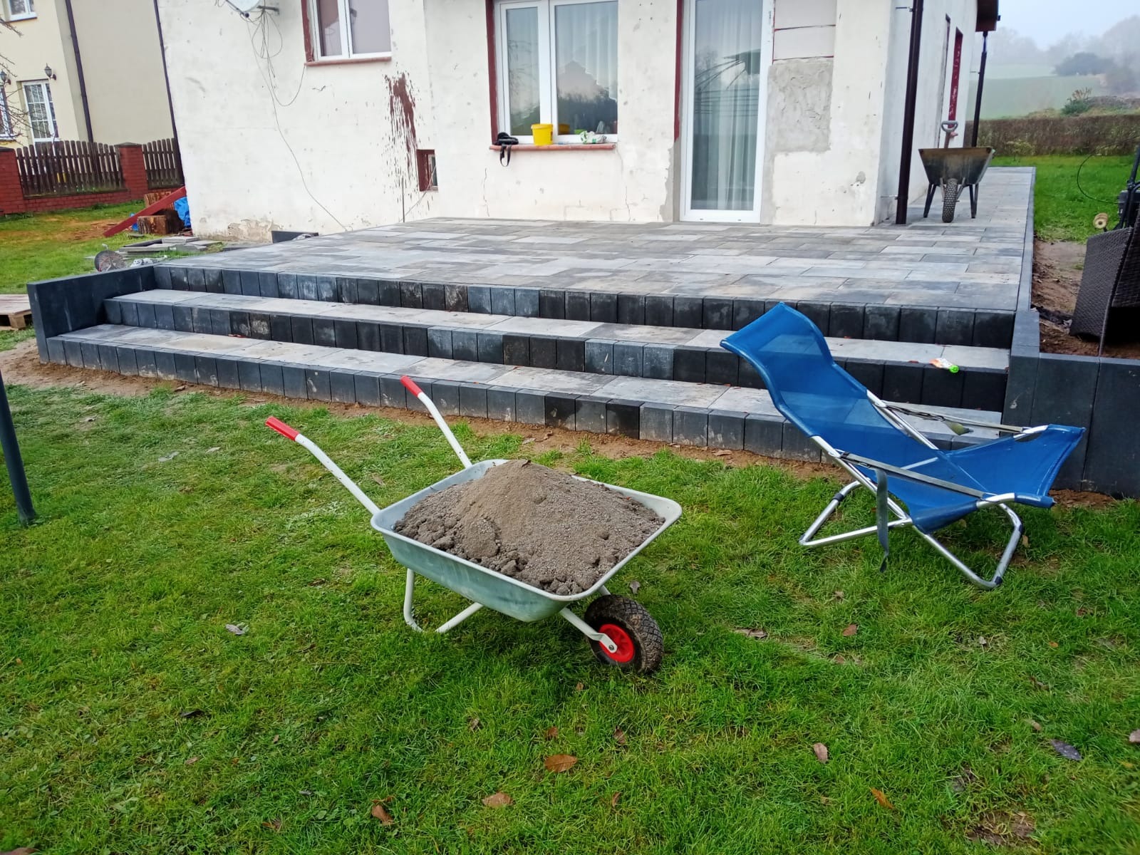 Nowo wybrukowany taras z ciemnoszarej kostki, schody z kostki, taczka z piaskiem i niebieski leżak na trawniku przed budynkiem.