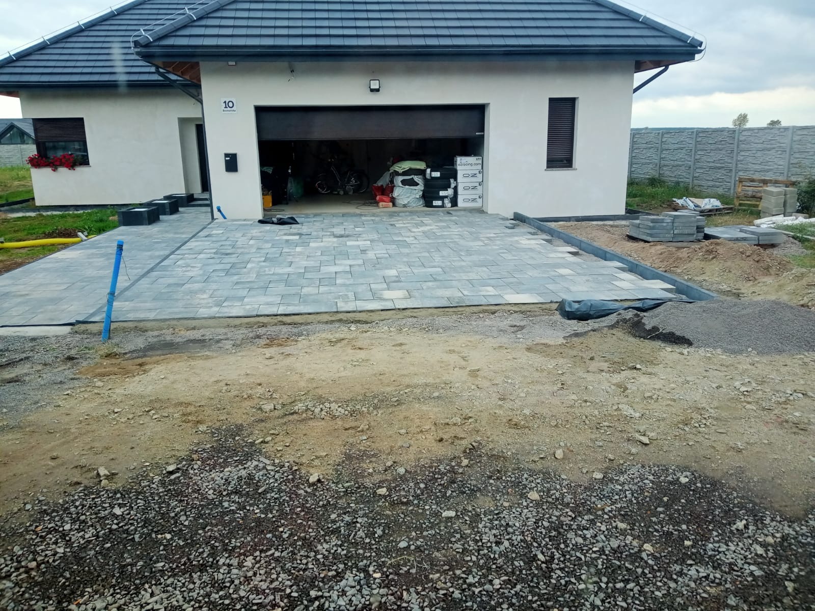 Ułożona szara kostka brukowa na podjeździe przed domem z otwartym garażem, widoczne przygotowania do dalszych prac, obrzeża, hałda kamieni i paleta z kostką w tle.