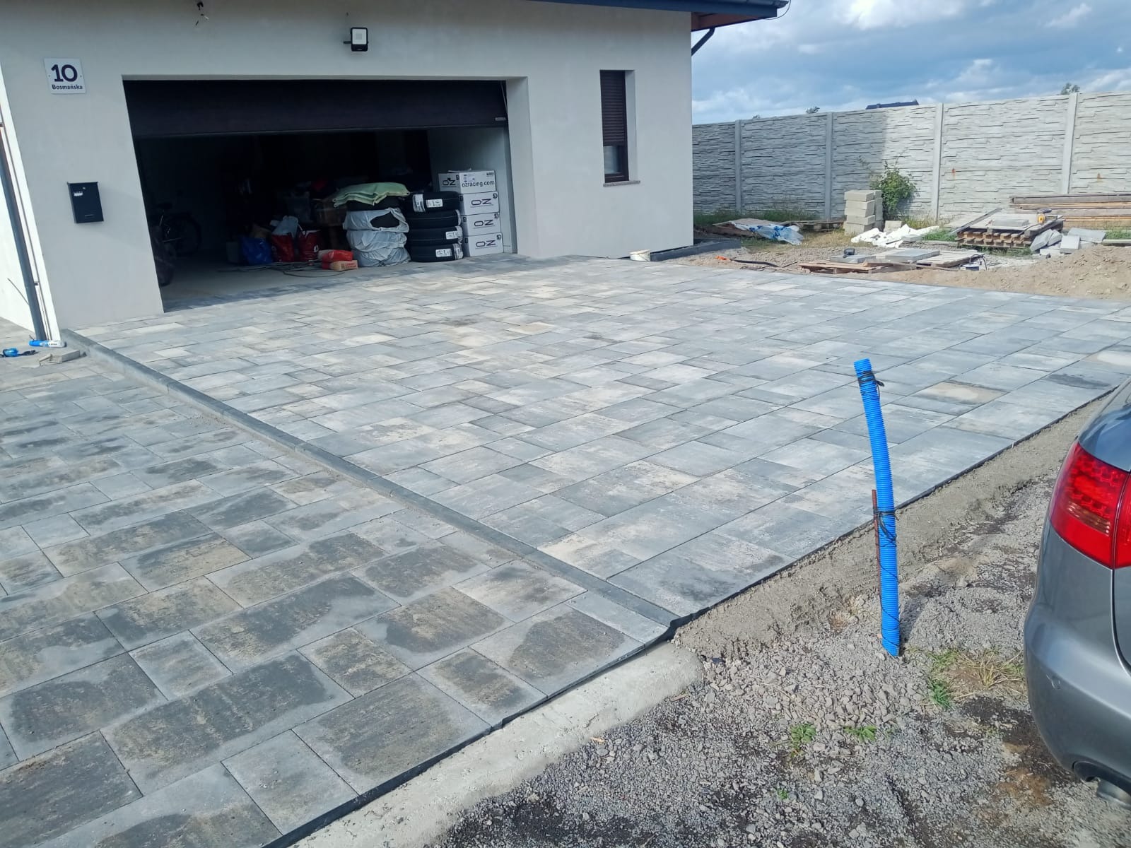 Nowo ułożony szary chodnik z kostki brukowej prowadzący do garażu, widoczny fragment betonowego ogrodzenia w tle, niebieska rura wystająca z ziemi obok zaparkowanego samochodu.
