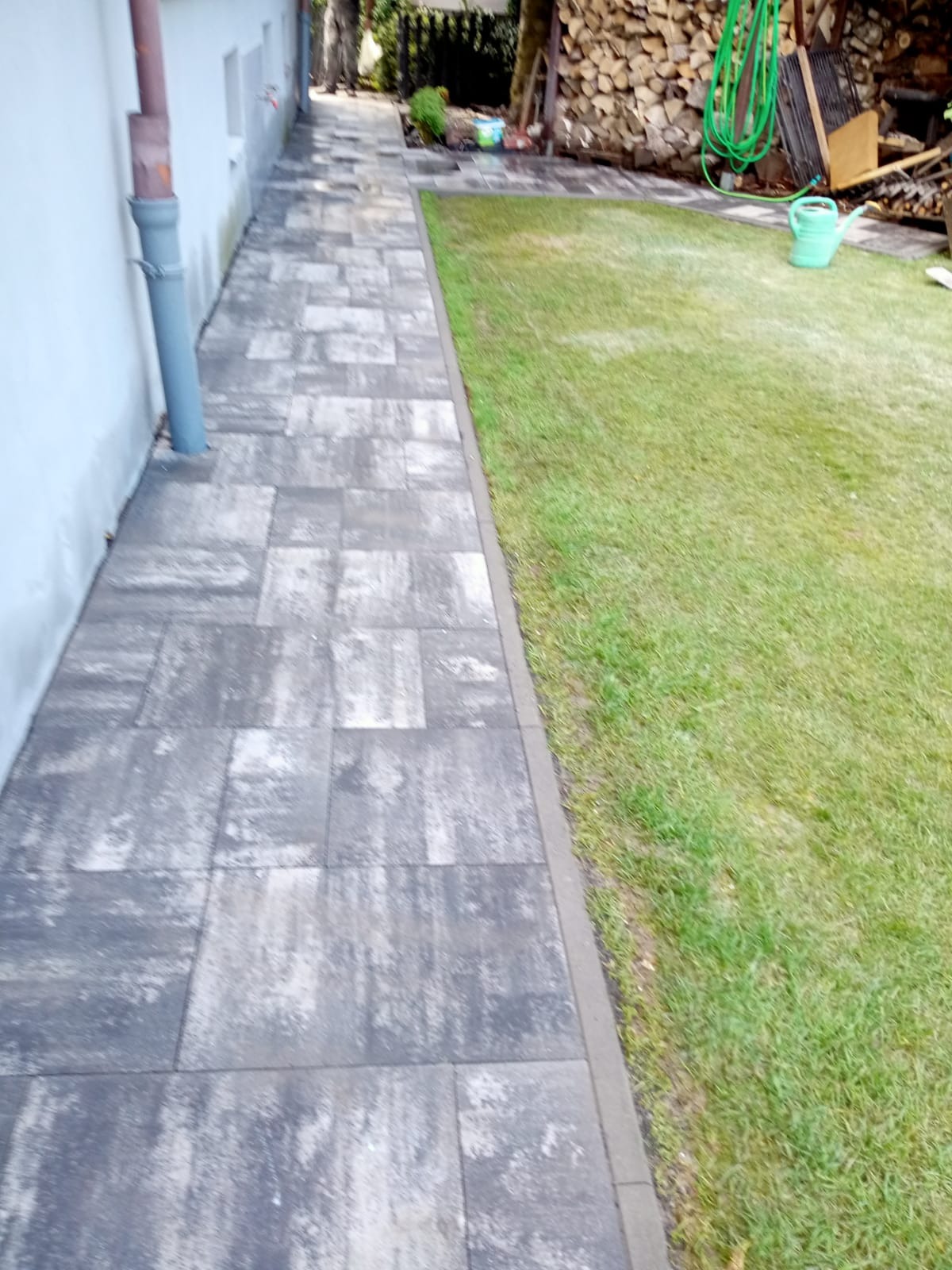 Ułożona ścieżka z szarej kostki brukowej prowadząca wzdłuż białej ściany budynku i trawnika. W tle widoczne drewno na opał i zielony wąż ogrodowy.