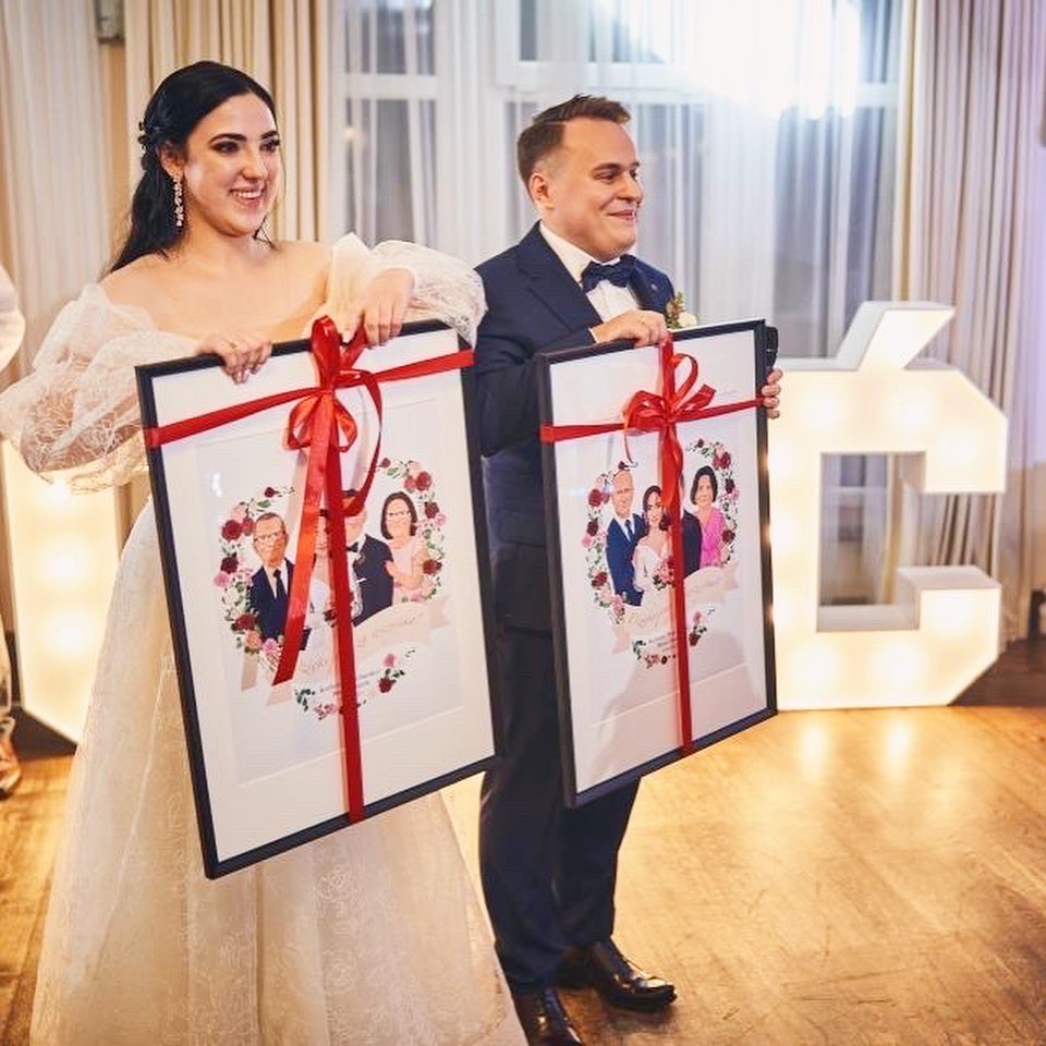 Para młoda prezentuje oprawione, spersonalizowane portrety rodzinne w formie ilustracji, przewiązane czerwoną wstążką, z dekoracyjnym napisem na dole, na tle sali weselnej z podświetlaną literą.