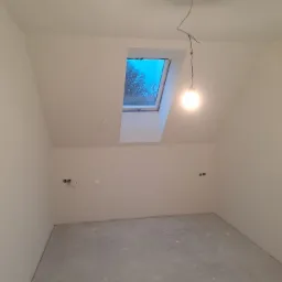 Poddasze po remoncie: świeżo pomalowane ściany w odcieniach bieli, surowy beton na podłodze, okno dachowe z widokiem na drzewa, wisząca żarówka na kablu i przygotowane gniazdka elektryczne.