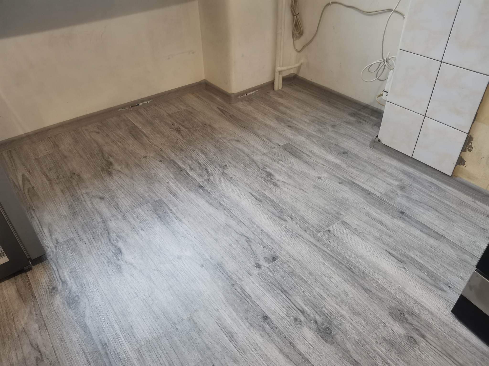 Ułożona podłoga z paneli imitujących jasne drewno w pomieszczeniu z białymi ścianami i widocznymi rurami grzewczymi.