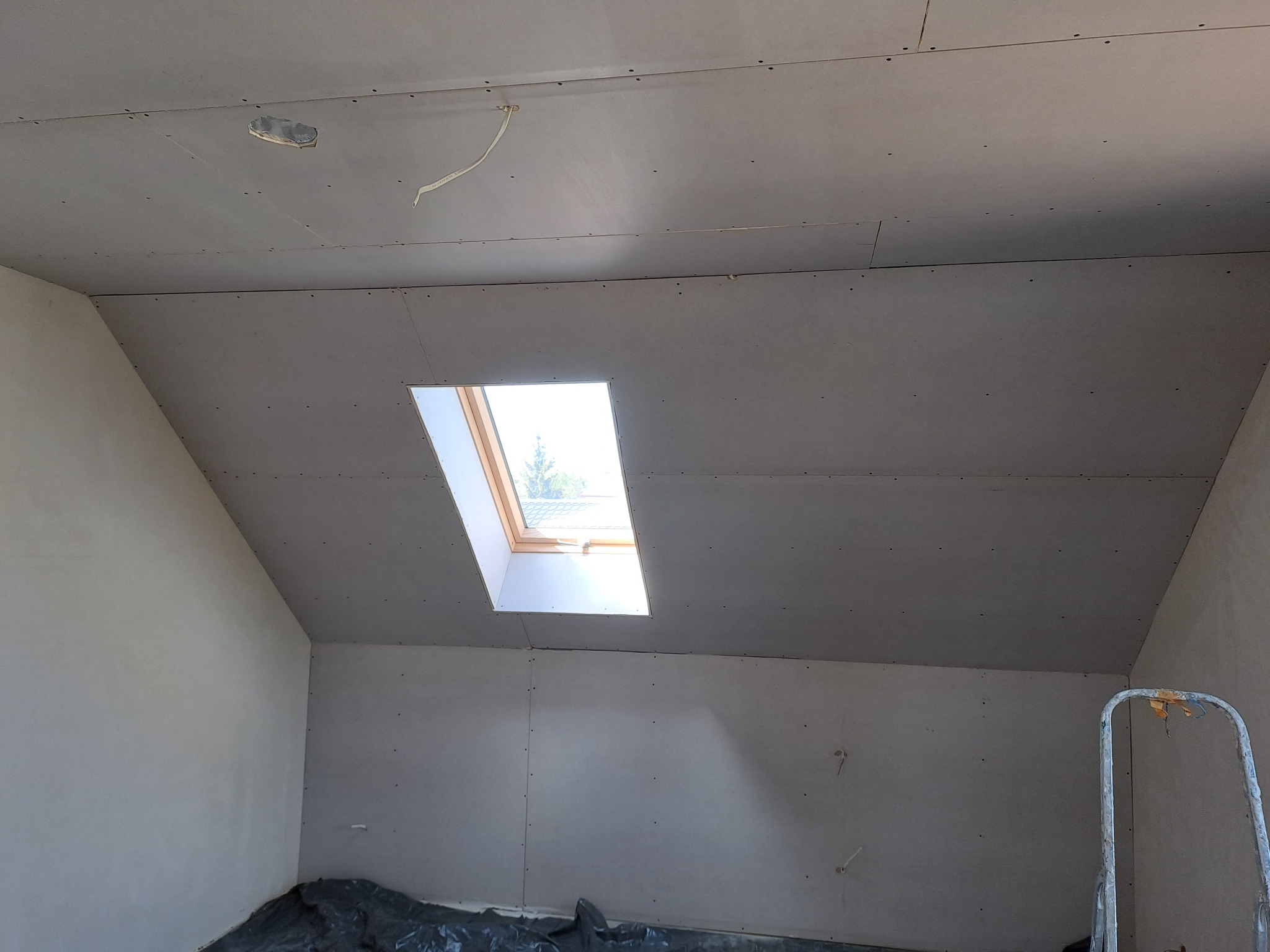 Poddasze z oknem dachowym w trakcie wykańczania płytami gipsowo-kartonowymi, widoczne kable elektryczne wystające z sufitu i drabina w rogu pomieszczenia.