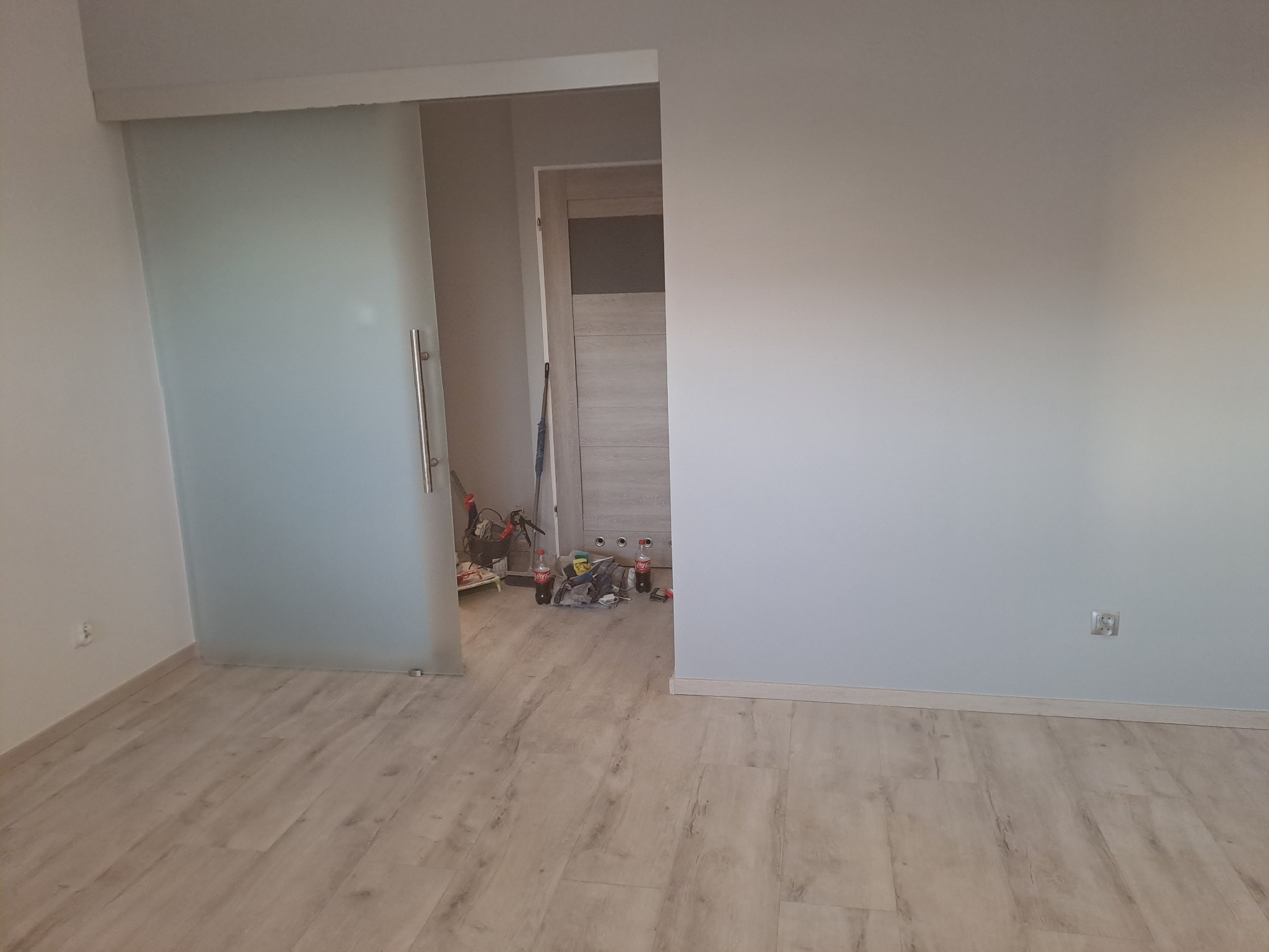 Wnętrze pokoju z przesuwnymi, szklanymi drzwiami; ściany pomalowane na jasny odcień, na podłodze panele imitujące drewno, widoczne gniazdko elektryczne na ścianie oraz narzędzia i materiały...