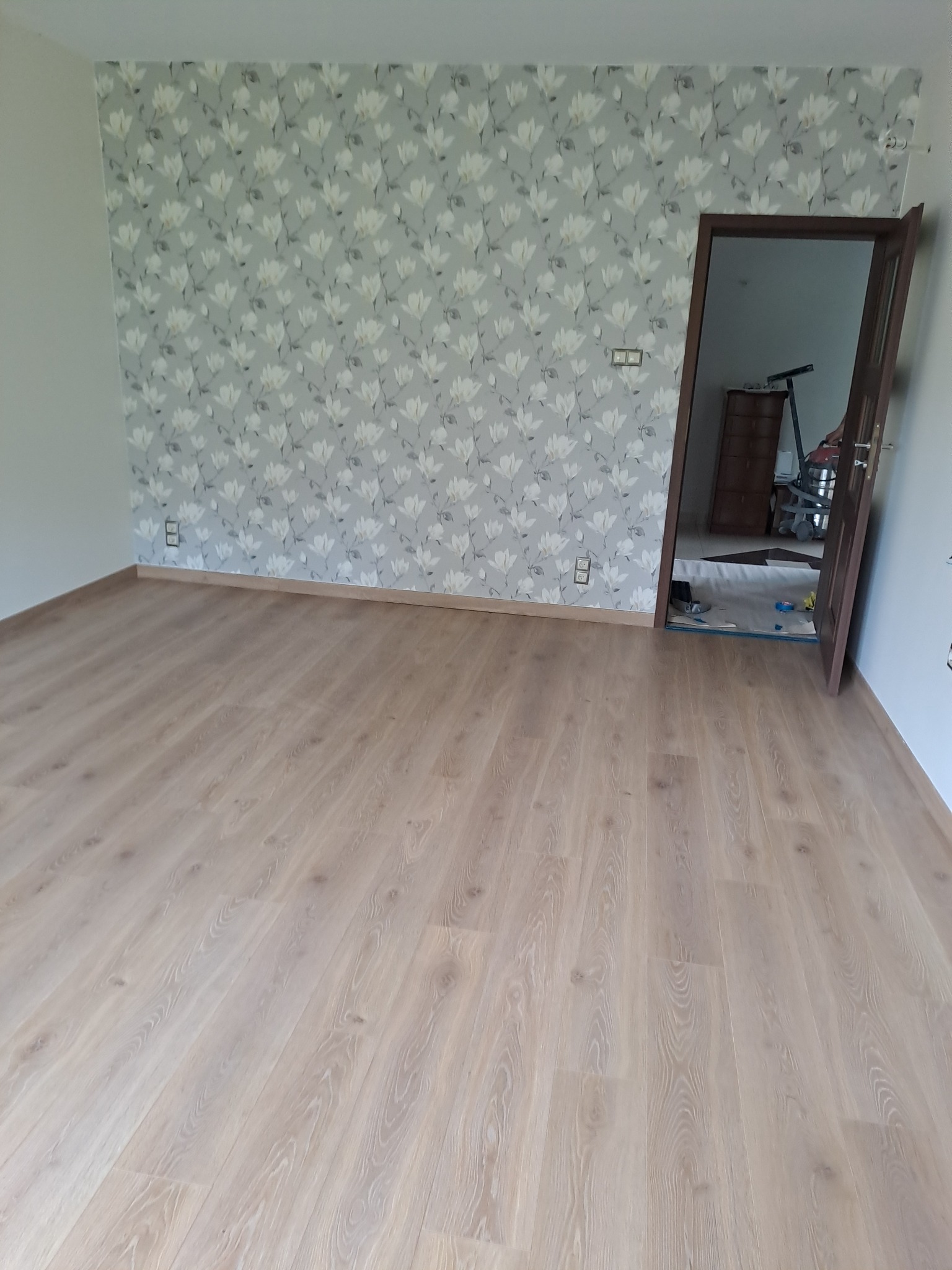 Pomalowane pomieszczenie z jasną podłogą i ścianą w kwiatowy wzór, widoczne gniazdka elektryczne i otwarte drzwi do innego pokoju z odkurzaczem.