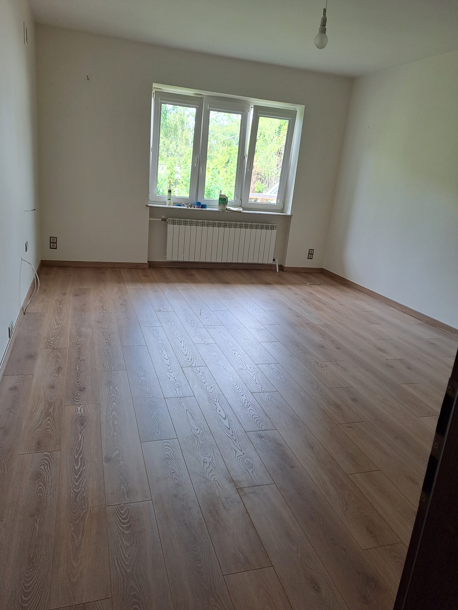 Pusty pokój po remoncie z panelami podłogowymi w odcieniach brązu, białymi ścianami z widocznymi gniazdkami elektrycznymi i oknem z białymi ramami wychodzącym na zieleń.