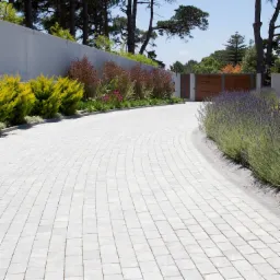 Zakrzywiony podjazd z jasnoszarej kostki brukowej, otoczony zielenią i prowadzący do drewnianej bramy w tle.