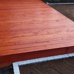 Kraina ogrodow - Uliana Kozak, Rostjslav Holodniak Spółka cywilna - Nowoczesny taras z drewna w odcieniach brązu, otoczony szarymi kostkami brukowymi, z widocznym fragmentem ogrodu i drzewami w tle.