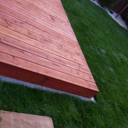 Kraina ogrodow - Uliana Kozak, Rostjslav Holodniak Spółka cywilna - Nowoczesny taras z desek w ciepłym odcieniu brązu, przylegający do zadbanego trawnika i fragmentu mebli ogrodowych.