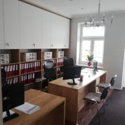 Biuro z drewnianymi blatami biurek i szafkami na dokumenty pod sufitem, oświetlone wiszącym żyrandolem, z widokiem na okno