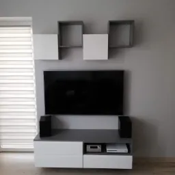 Nowoczesna szafka RTV z białymi frontami i szarym blatem, uzupełniona o wiszące, geometryczne półki w kolorach bieli i szarości, z telewizorem i głośnikami, na tle szarej ściany i drewnianej podłogi.