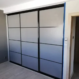 Trzydrzwiowa szafa wnękowa z przesuwnymi, matowymi drzwiami w kolorze szaro-niebieskim, podzielonymi czarnymi listwami na dziewięć prostokątnych pól, widoczna w rogu pokoju na tle szarej ściany...