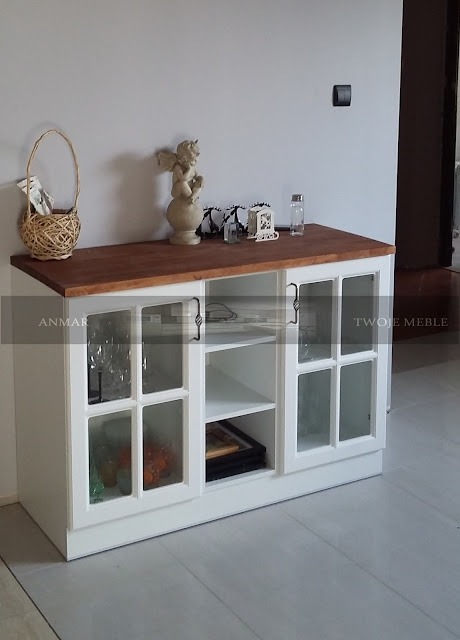 Biała komoda z przeszklonymi drzwiczkami w stylu rustykalnym, drewniany blat w ciepłym odcieniu brązu, dekoracje na blacie: wiklinowy koszyk, figurka aniołka i szklane pojemniki.