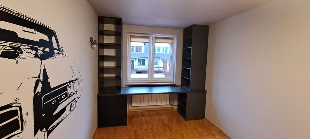 Szare biurko z regałami po bokach okna, na ścianie grafika z samochodem GTO, drewniana podłoga.