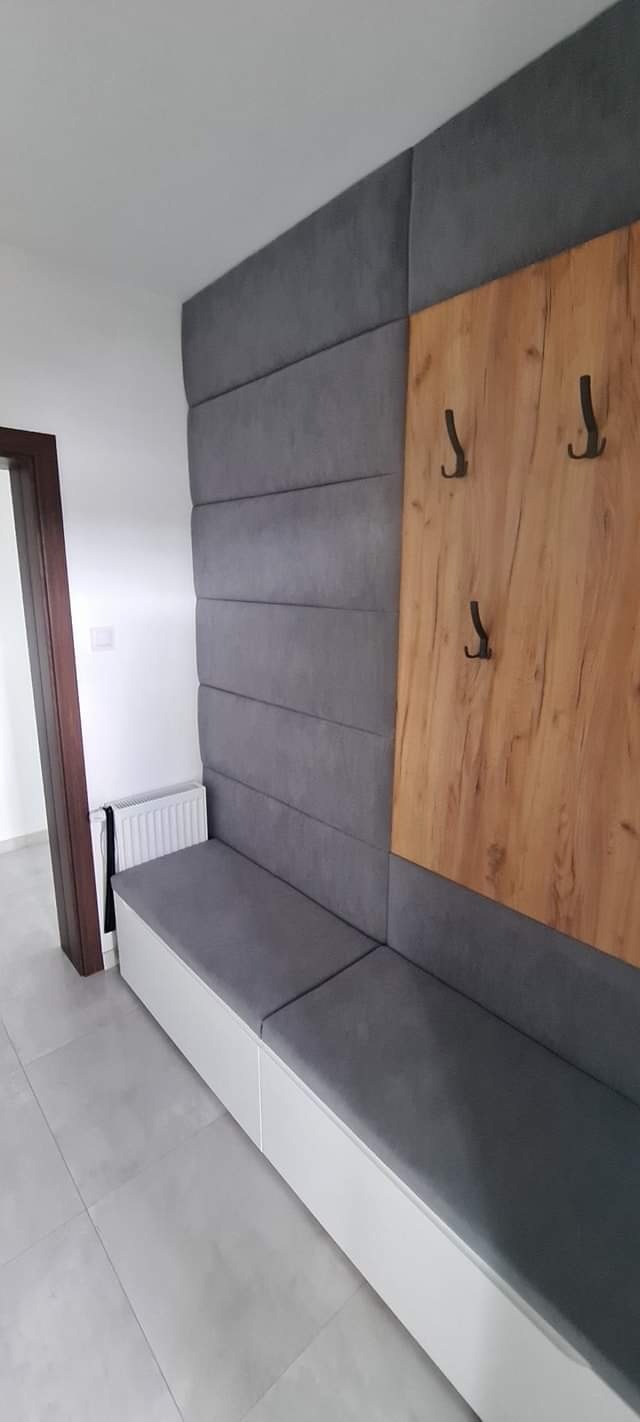 Panel tapicerowany w przedpokoju z siedziskiem i drewnianym panelem z wieszakami, widoczny grzejnik i fragment drzwi