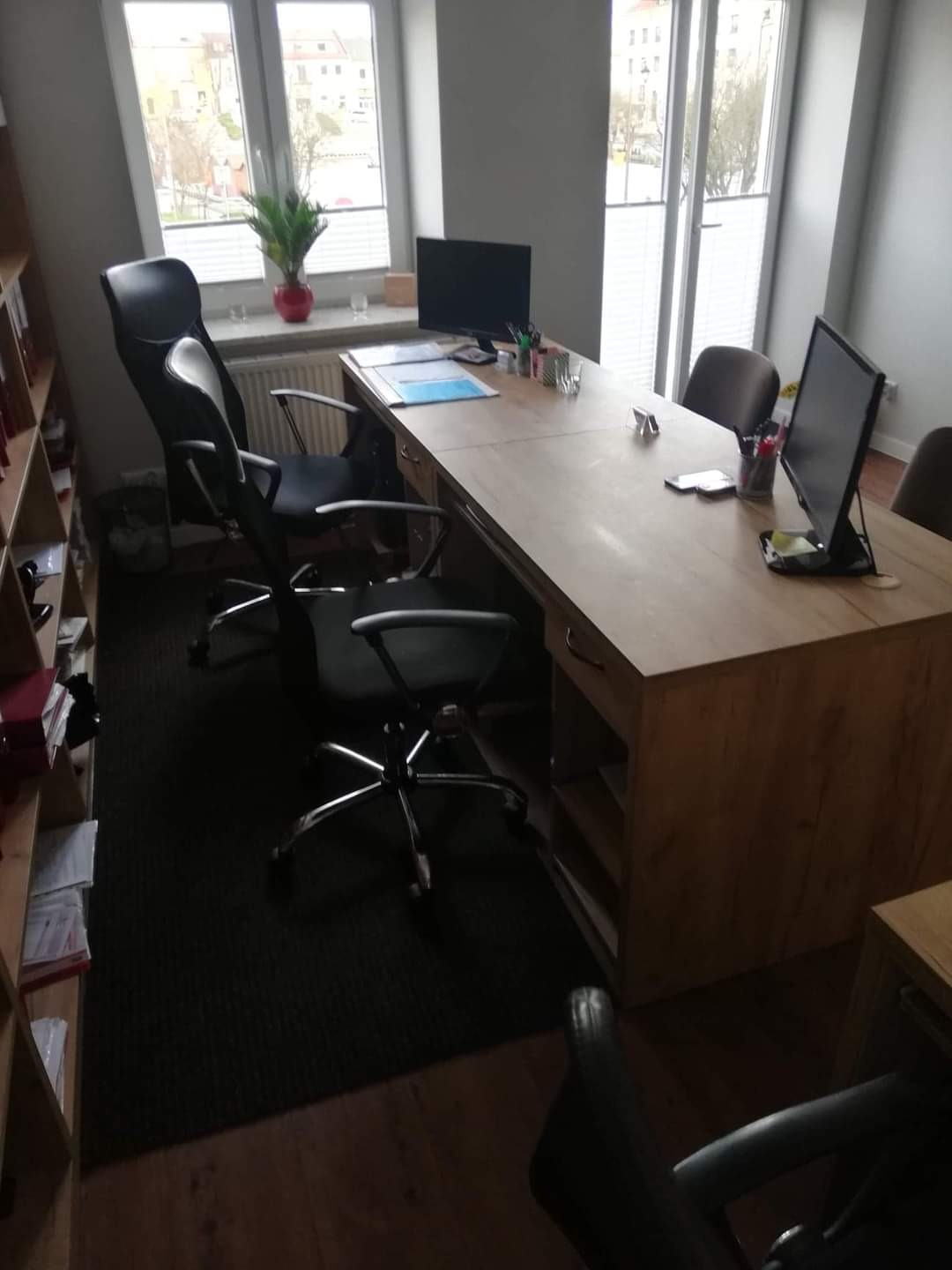 Duży, drewniany blat biurka w jasnym odcieniu drewna, z dwoma stanowiskami pracy, krzesłami obrotowymi i monitorami, umieszczony w biurze z oknami i regałem na dokumenty.