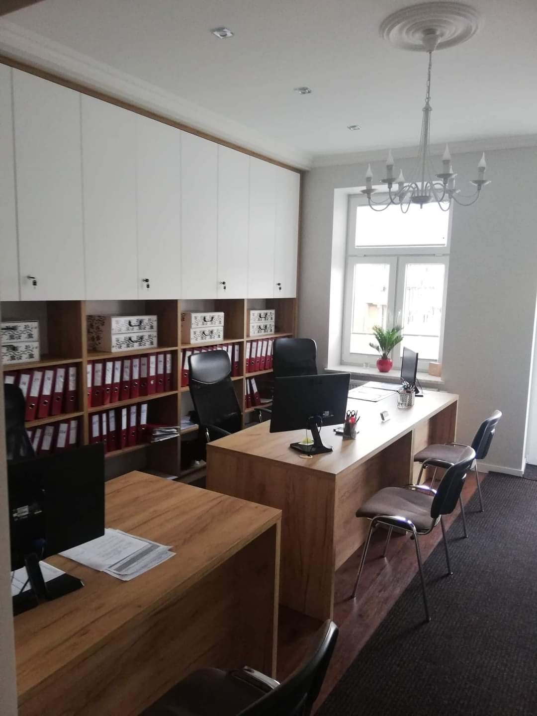 Biuro z drewnianymi blatami biurek i szafkami na dokumenty pod sufitem, oświetlone wiszącym żyrandolem, z widokiem na okno