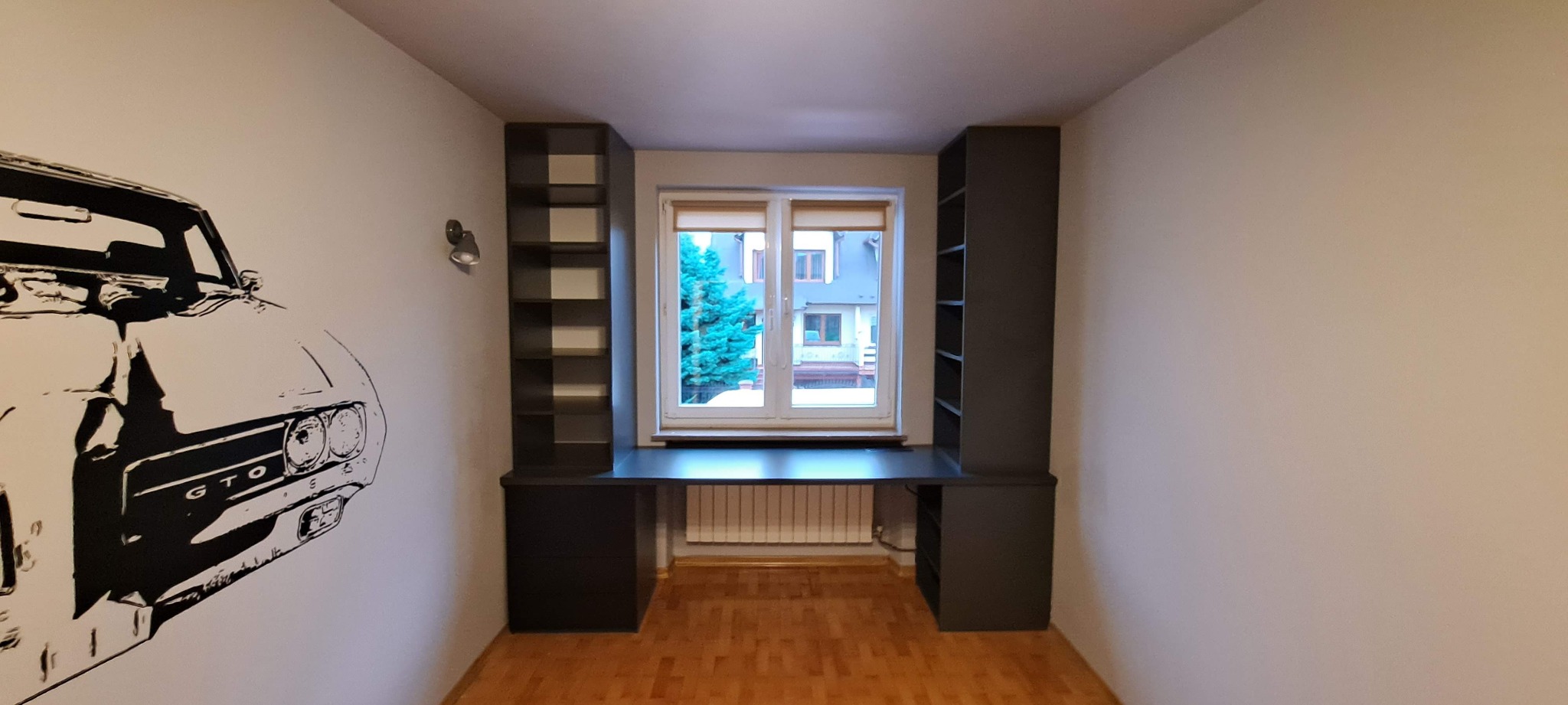 Pokój z szarą zabudową meblową wokół okna, tworzącą biurko z półkami, z grafiką samochodu GTO na ścianie, parkiet na podłodze.