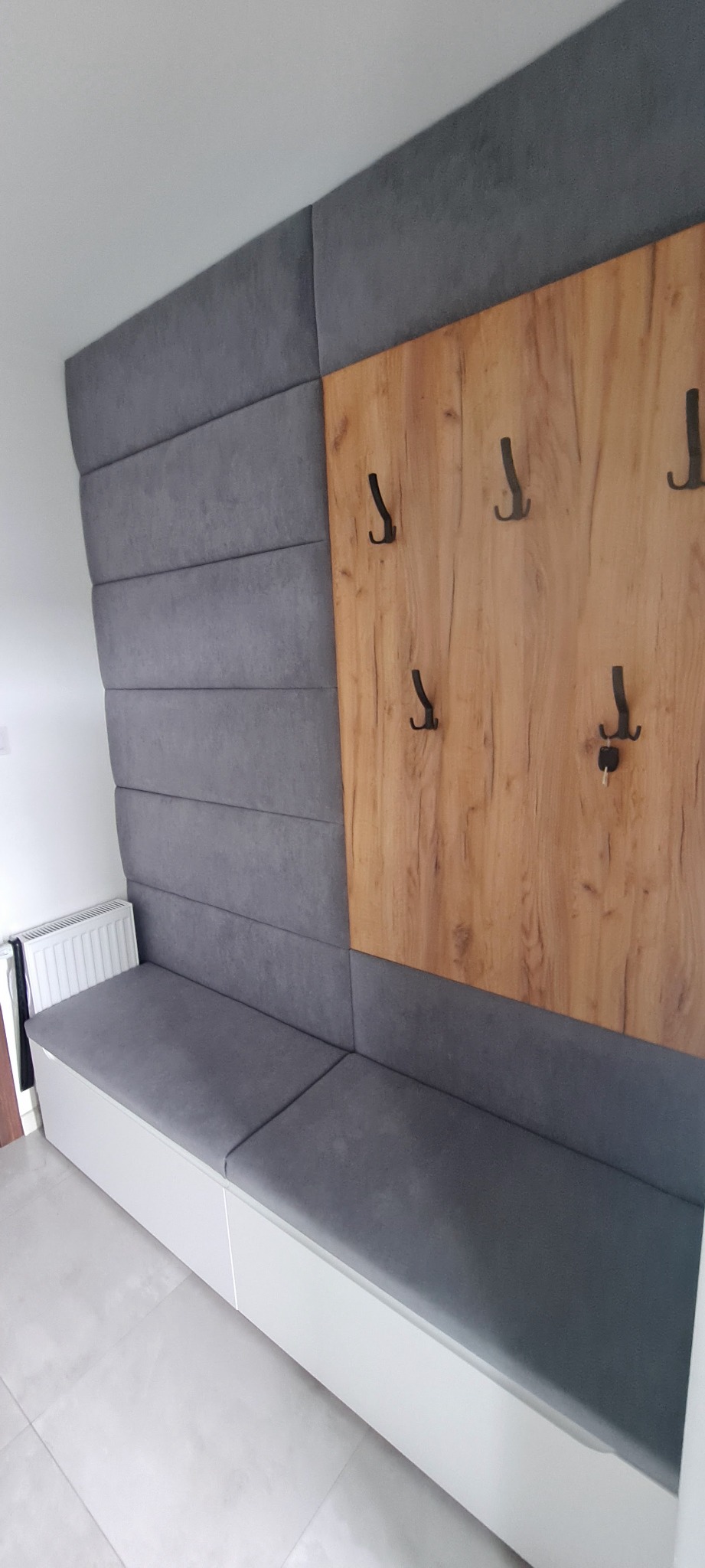 Szary panel tapicerowany z drewnianą wstawką z haczykami, tworzący siedzisko i wieszak w przedpokoju.