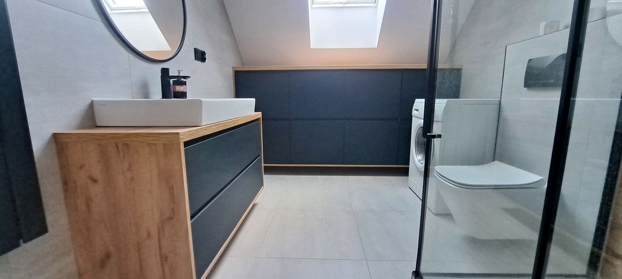 Nowoczesna łazienka na poddaszu z drewnianą szafką pod umywalką, zabudową w kolorze granatowym oraz toaletą za szklaną kabiną prysznicową.
