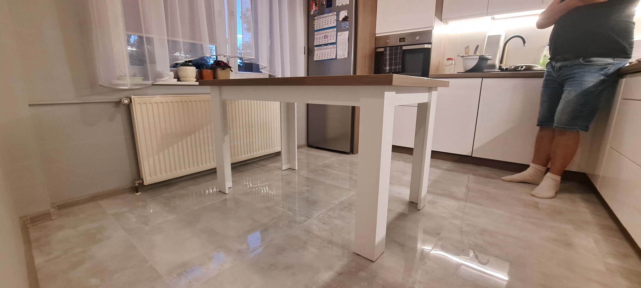 Stół z blatem w kolorze drewna i białymi nogami w nowoczesnej kuchni, widoczne białe szafki i fragment osoby w jeansowych szortach.