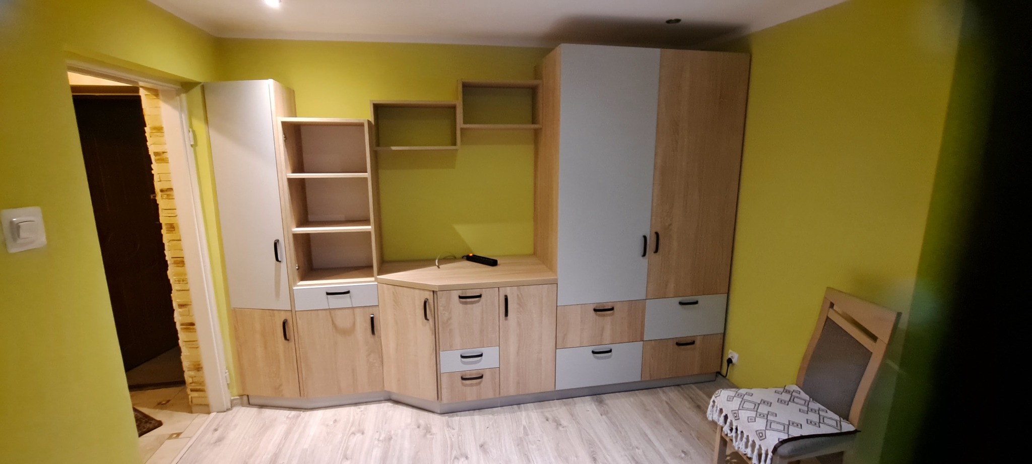 Zestaw mebli z drewna i szarego laminatu w pokoju z limonkowymi ścianami, obejmujący szafę, regał z półkami, szafki z szufladami i półkami, zwieńczony otwartymi półkami wiszącymi; krzesło...