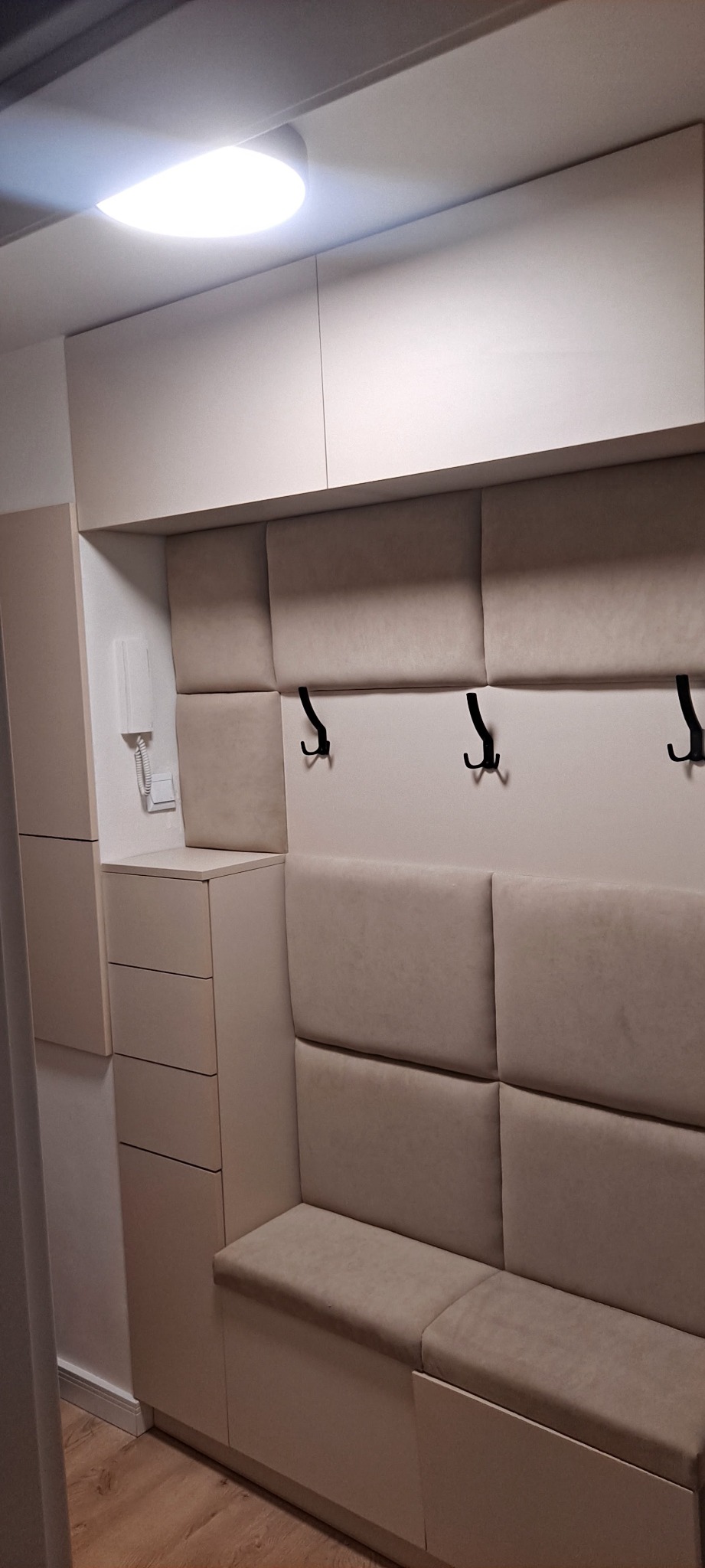 Beżowa zabudowa przedpokoju z siedziskiem, wieszakami i szafkami, oświetlona lampą sufitową.