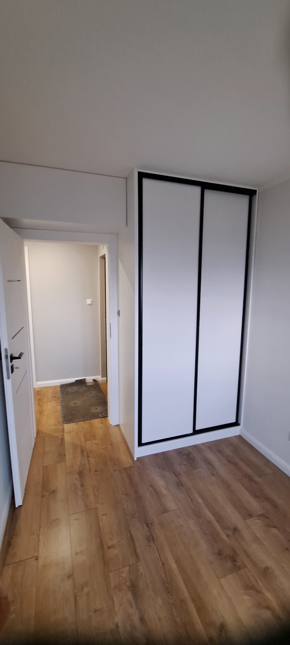 Biała szafa wnękowa z przesuwnymi drzwiami i czarnymi obramowaniami w jasnym pokoju z drewnianą podłogą, widok na otwarte drzwi do kolejnego pomieszczenia.