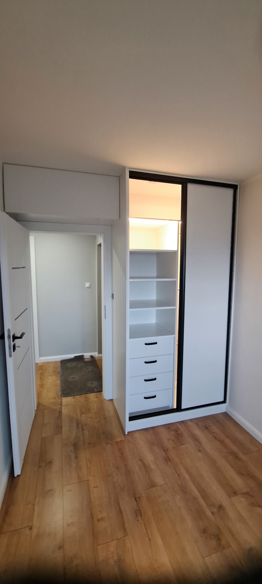 Biała szafa wnękowa z przesuwnymi drzwiami i czarnymi obramowaniami, z otwartą częścią z półkami i szufladami, oświetlona od wewnątrz, widoczna z perspektywy wnętrza mieszkania z drewnianą podłogą.