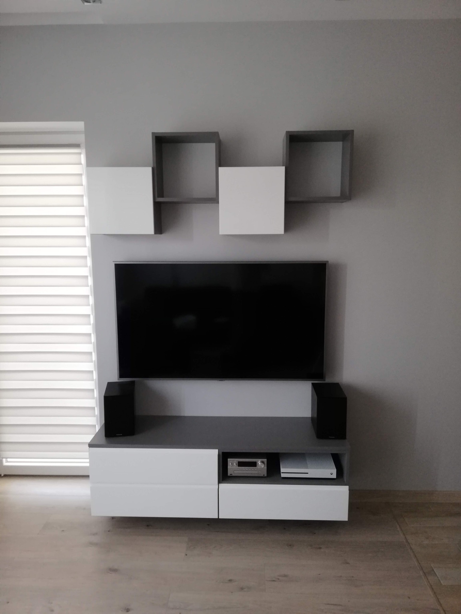 Nowoczesna szafka RTV z białymi frontami i szarym blatem, uzupełniona o wiszące, geometryczne półki w kolorach bieli i szarości, z telewizorem i głośnikami, na tle szarej ściany i drewnianej podłogi.