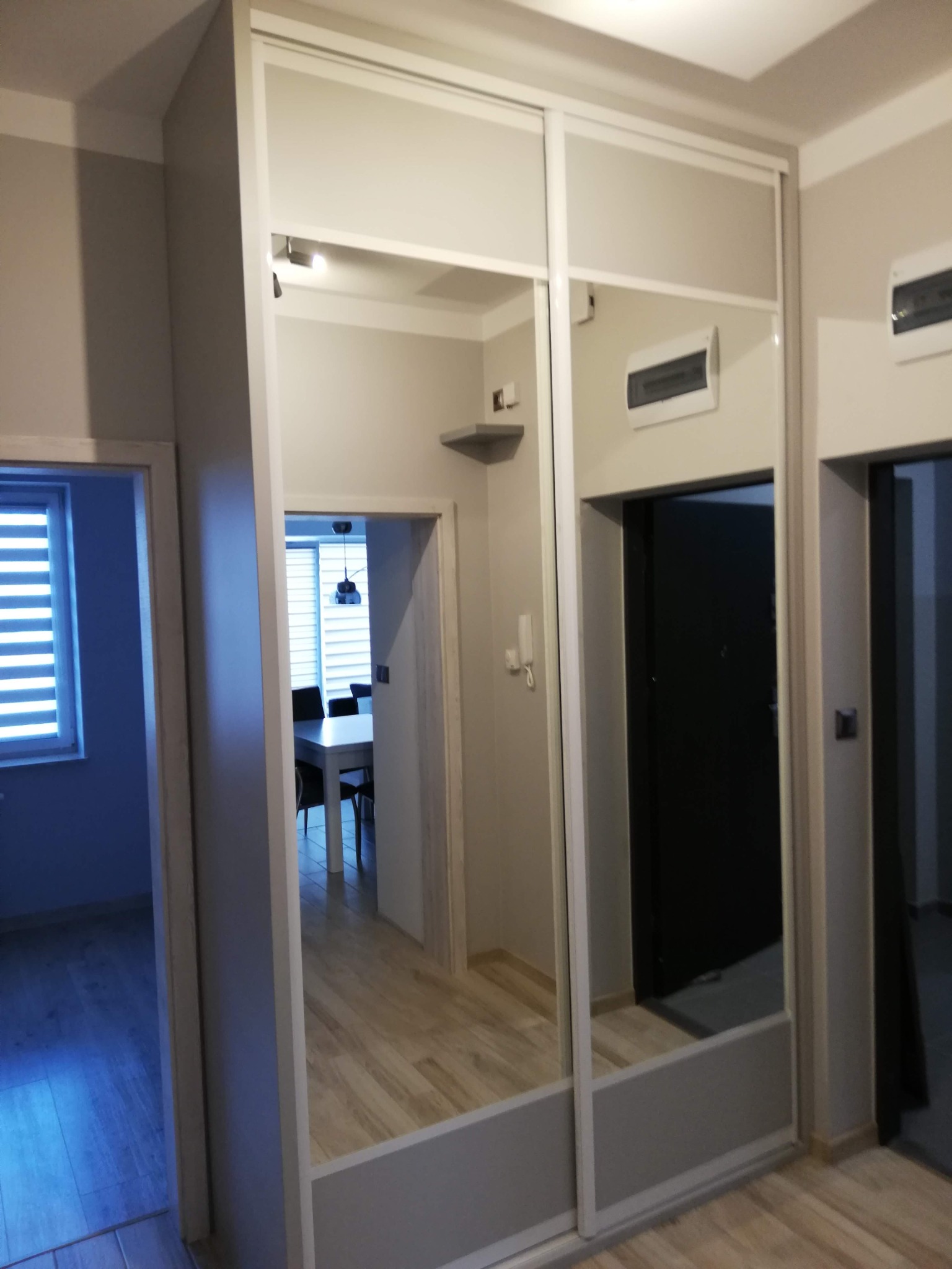 Biała szafa wnękowa z lustrzanymi drzwiami przesuwnymi, odbijająca fragment mieszkania z widoczną jadalnią i oknem z żaluzjami.