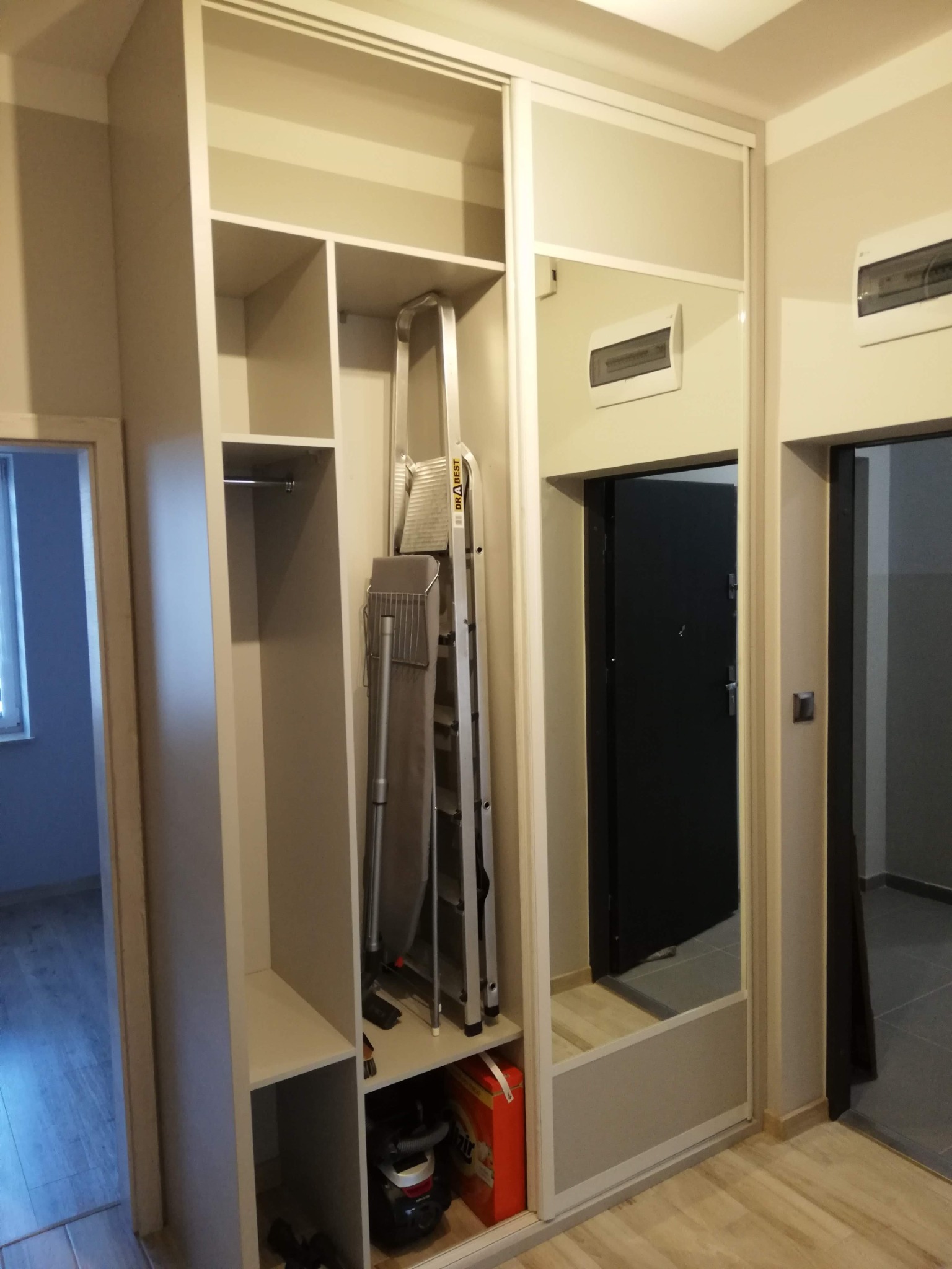 Jasna szafa wnękowa z przesuwnymi drzwiami lustrzanymi, z widocznym wnętrzem zawierającym półki, drążek na ubrania, deskę do prasowania i drabinę, z odkurzaczem w dolnej części.