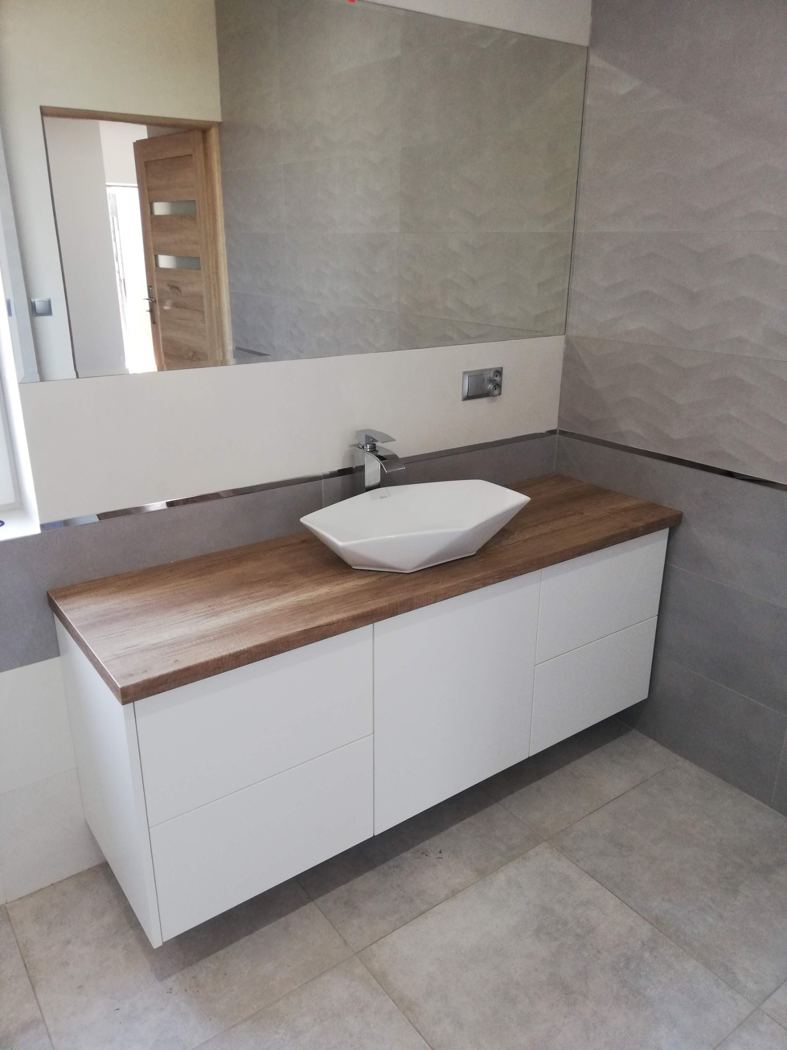 Nowoczesna łazienka z drewnianym blatem, białą szafką pod umywalkę w kształcie rombu i dużym lustrem z widocznym odbiciem drzwi.